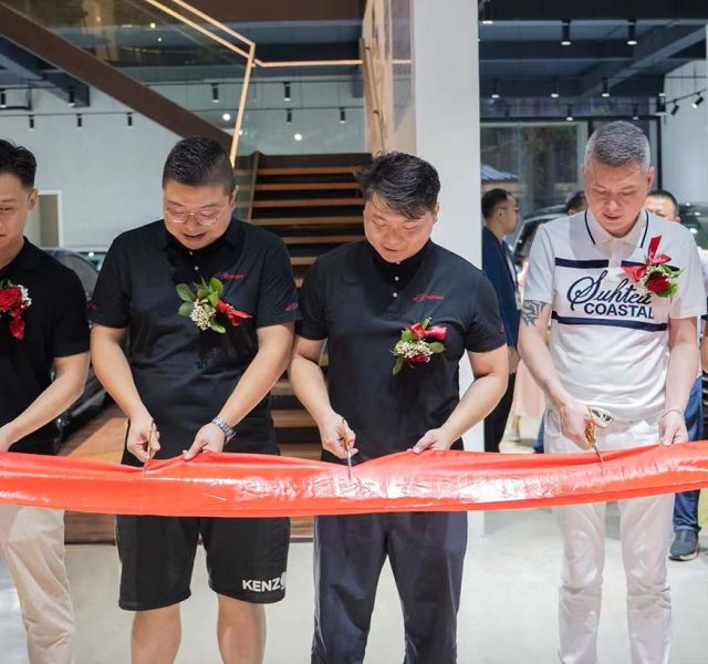 A group of people participating in a ribbon - cutting ceremony, likely for a business opening, with a red ribbon and floral decorations.