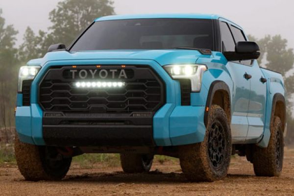 Turquoise Toyota truck (front view) on muddy dirt, "TOYOTA" grille, fog lights on, wooded backdrop.