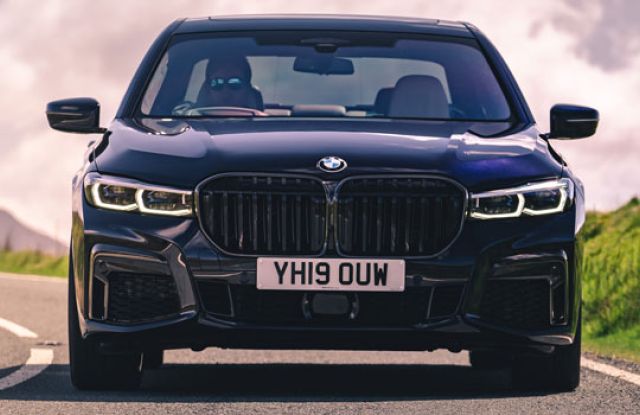 Front view of a dark BMW sedan on a rural road: Features a prominent grille, glowing LED headlights, and a white license plate, emphasizing its luxury design against a scenic, cloudy backdrop.