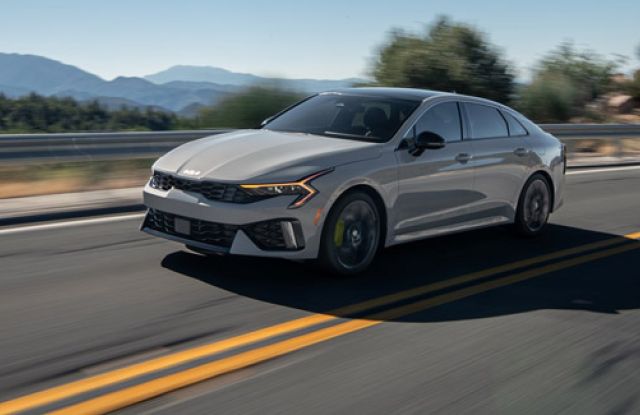 Kia K5 2025 sedan driving on highway (motion blur), mountain/wooded backdrop, yellow road lines.