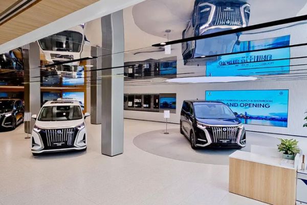 A modern car showroom featuring three luxury MPVs, with sleek designs, large digital screens announcing a grand opening, and a stylish, reflective interior with plants and wooden accents.