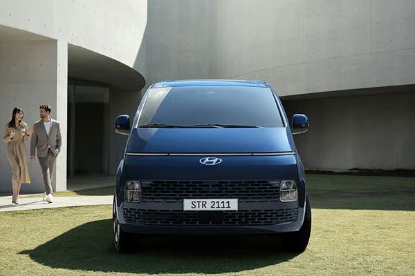 A blue Hyundai minivan with the license plate "STR 2111" parked on a grassy area, with two formally dressed people walking nearby and a modern concrete building in the background.