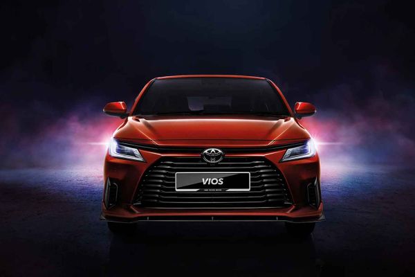 A red Toyota Vios sedan showcased in a dramatic, dark studio setting with colorful lighting effects, emphasizing its bold front design and modern aesthetic.