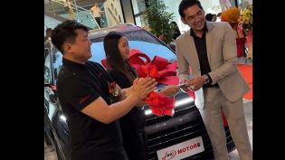 People at a car dealership ceremony, with one person in a suit handing over something to another in front of a car adorned with a big red bow.