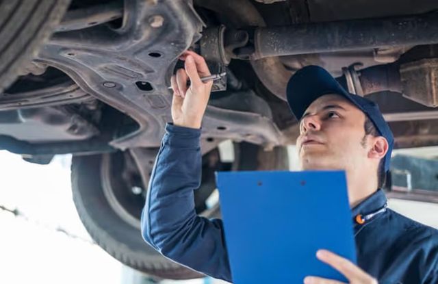 A mechanic in a blue uniform and cap inspects a car's undercarriage with a tool, holding a blue clipboard, in an auto repair shop.