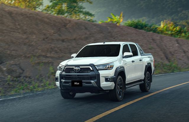 A white Toyota Hilux pickup truck driving on a road, with a rugged, green-hilled landscape in the background, showcasing its robust design.