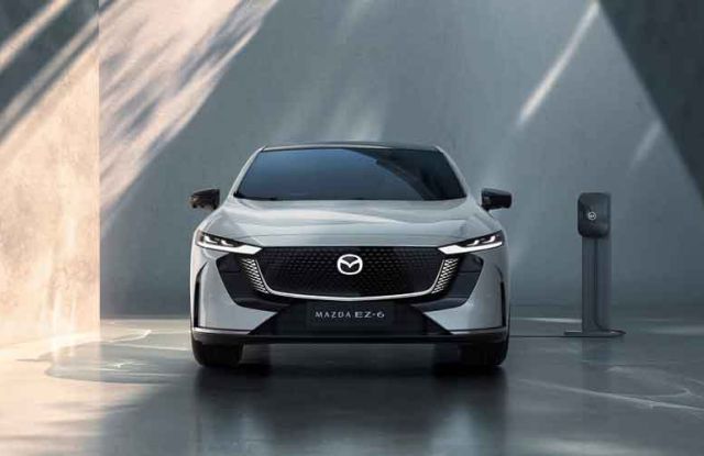 Front view of a silver Mazda EZ-6 electric sedan, charging at a station in a minimalist, futuristic space with dramatic light and shadow on concrete walls.