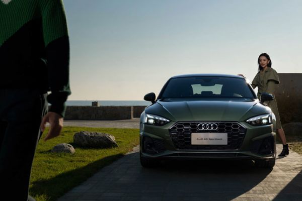 Green Audi A5 Sportback (front view): Features Audi logo and badging, parked by a coastal area with two people nearby—showcasing its sleek, premium design in an outdoor setting.
