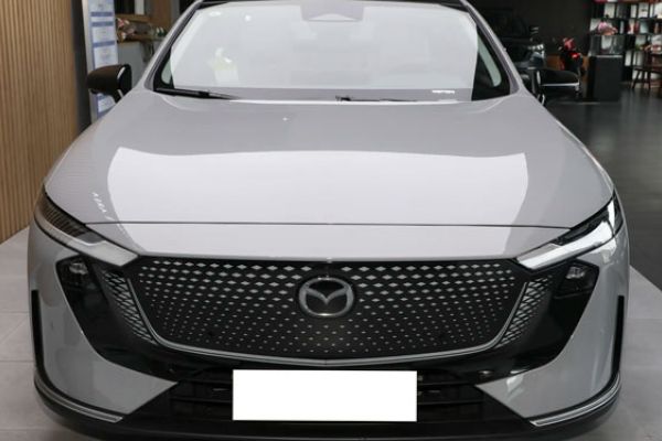 Front view of a light gray Mazda sedan (modern design) in a showroom: Features a textured grille with Mazda logo, slim LED headlights, and sleek black accents. Showroom interior (furniture, displays) visible in the background.