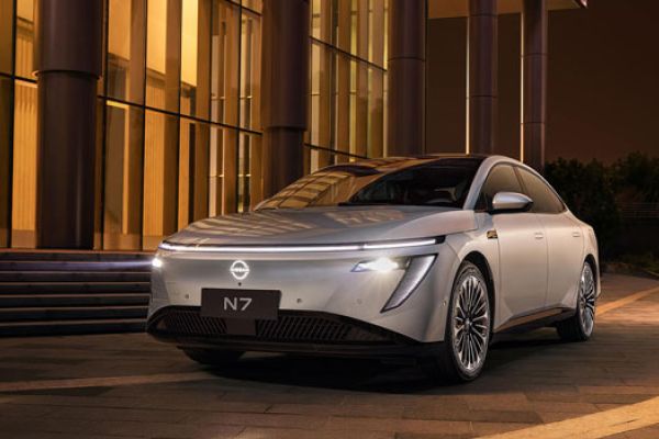 Silver N7 electric sedan parked at night outside a modern building (lit glass walls, columns). The sleek car’s illuminated headlights and "N7" badge are visible; warm lighting and paved ground enhance its contemporary design.