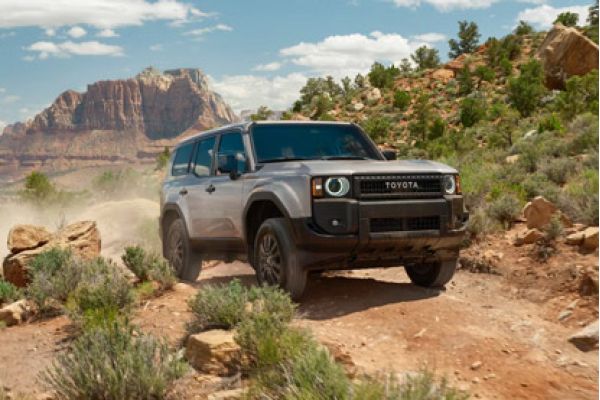 Silver 2026 Toyota Land Cruiser driving on a rugged desert trail, with a dramatic red rock mountain backdrop.