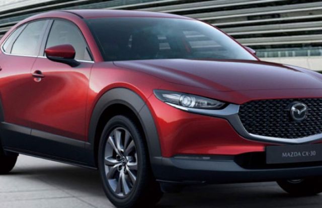 Red Mazda CX-30 crossover: Features Mazda logo, "MAZDA CX-30" badging, sleek headlights, and dual-tone alloy wheels—displayed by a modern building, showcasing its sporty design.