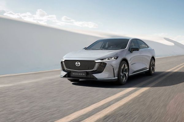 A silver Mazda EZ-6 electric sedan driving on a road, with a backdrop of white sand dunes and a clear blue sky.