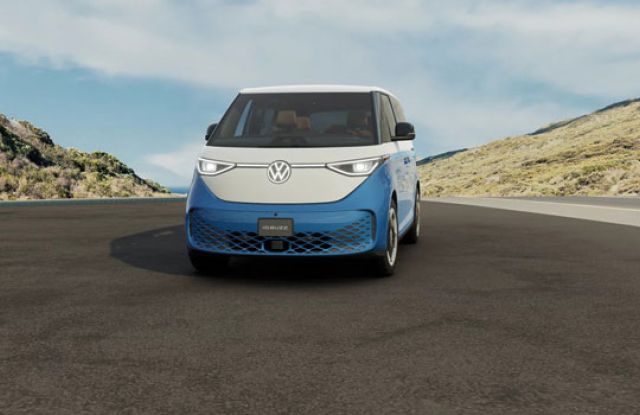 Front view of a 2025 Volkswagen ID. Buzz on a road, set against hilly, vegetated landscape under a partly cloudy sky.