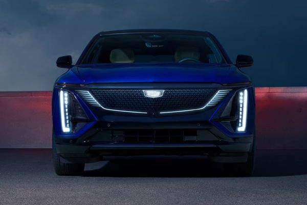 Glossy blue Cadillac Lyriq electric SUV, showcased with its illuminated signature headlights against a moody, lit backdrop.