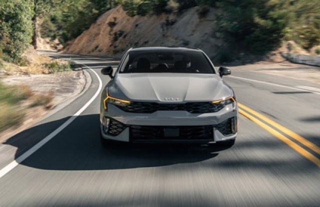 Kia K5 2025 (front view) driving on winding mountain road (motion blur), rugged landscape backdrop.