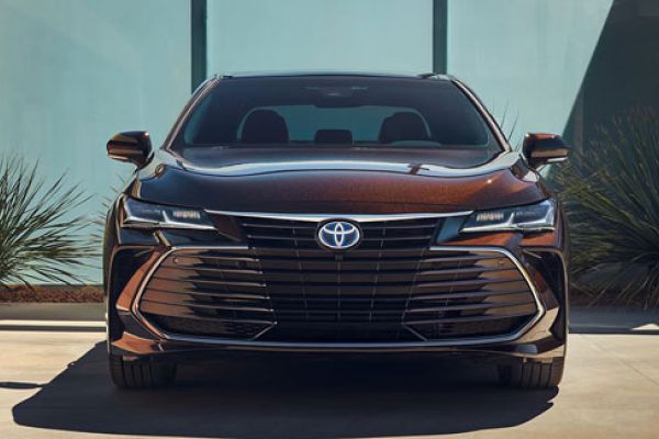 Front view of a dark bronze Toyota sedan: Features a prominent Toyota logo, sleek LED headlights, and a modern grille. Set against a contemporary building/desert plant backdrop, highlighting its upscale design.