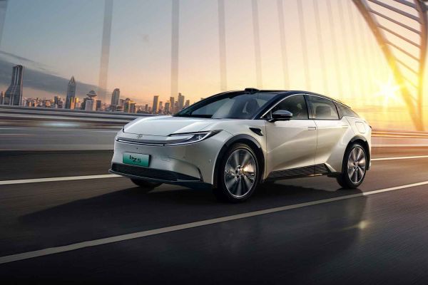 A white Toyota bZ5 electric car driving on a bridge at sunset, with a city skyline in the background, showcasing its futuristic design and dynamic driving scene.