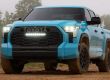 Turquoise Toyota truck (front view) on muddy dirt, "TOYOTA" grille, fog lights on, wooded backdrop.