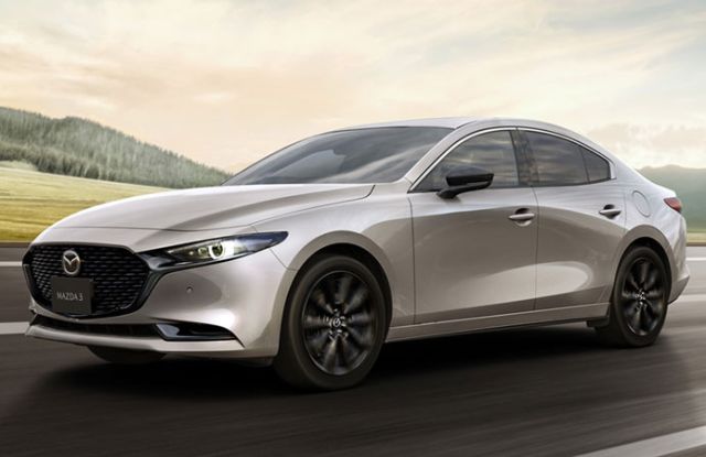 A silver Mazda3 sedan driving on a road with a scenic landscape of hills and fields, highlighting its sleek design and dynamic performance.