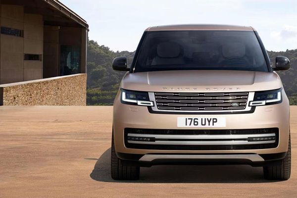 A gold Range Rover SUV with "176 UYP" license plate, parked near a modern building with a scenic mountain background, highlighting its luxury and bold front design.