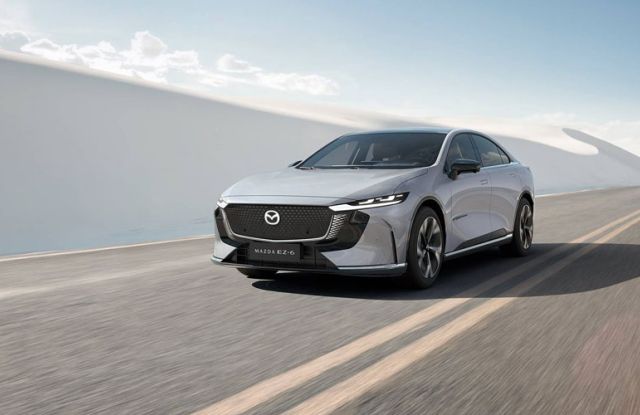 A silver Mazda EZ-6 electric sedan driving on a road, with a backdrop of white sand dunes and a clear blue sky.