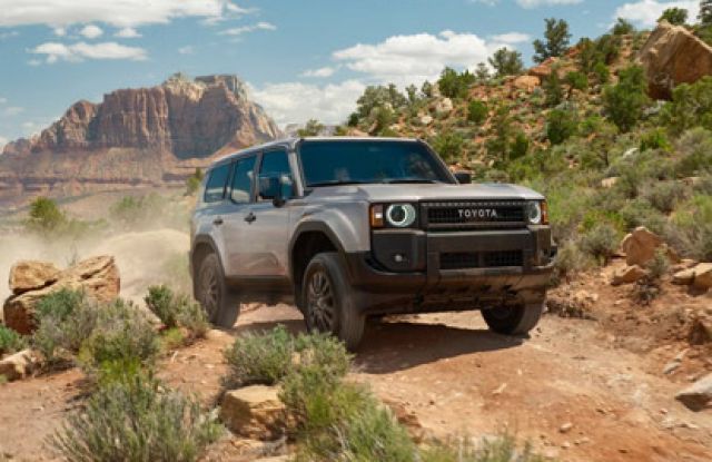 Silver 2026 Toyota Land Cruiser driving on a rugged desert trail, with a dramatic red rock mountain backdrop.