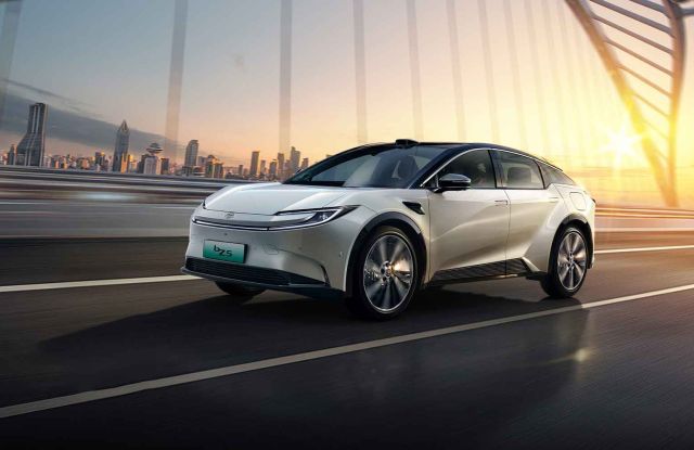 A white Toyota bZ5 electric car driving on a bridge at sunset, with a city skyline in the background, showcasing its futuristic design and dynamic driving scene.