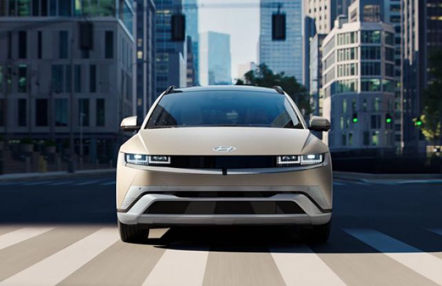 Beige Hyundai Ioniq 5 crossing a city crosswalk with tall modern buildings in the background.