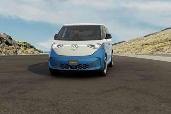 Front view of a 2025 Volkswagen ID. Buzz on a road, set against hilly, vegetated landscape under a partly cloudy sky.