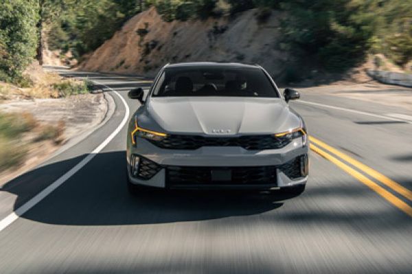 Kia K5 2025 (front view) driving on winding mountain road (motion blur), rugged landscape backdrop.