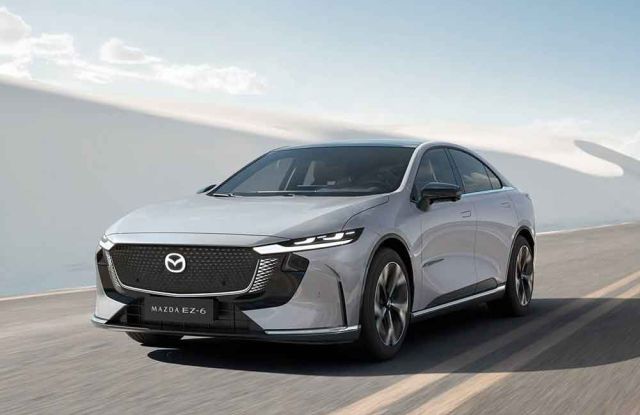 A silver Mazda EZ-6 sedan driving on a road, with a vast, light-colored landscape and clear blue sky in the background, showcasing its sleek and modern design.