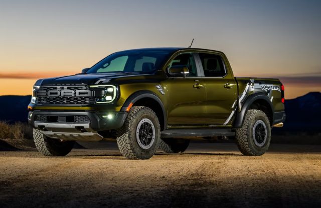 A green Ford Ranger Raptor pickup truck parked on a dirt road, with a scenic sunset and mountainous landscape in the background.