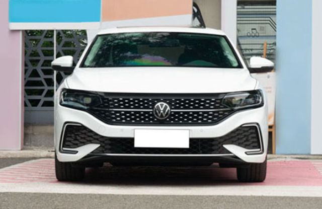 White Volkswagen sedan (direct front view): features a black patterned grille (VW logo), sleek headlights, and sporty lower vents—parked by a colorful structure, showcasing its modern bold design.