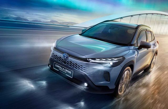 A silver Toyota Corolla Cross Hybrid Electric SUV driving on a road, with a scenic background featuring a bridge and a colorful sky.