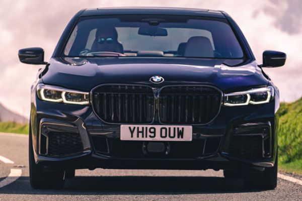 Front view of a dark BMW sedan on a rural road: Features a prominent grille, glowing LED headlights, and a white license plate, emphasizing its luxury design against a scenic, cloudy backdrop.