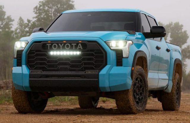Turquoise Toyota truck (front view) on muddy dirt, "TOYOTA" grille, fog lights on, wooded backdrop.