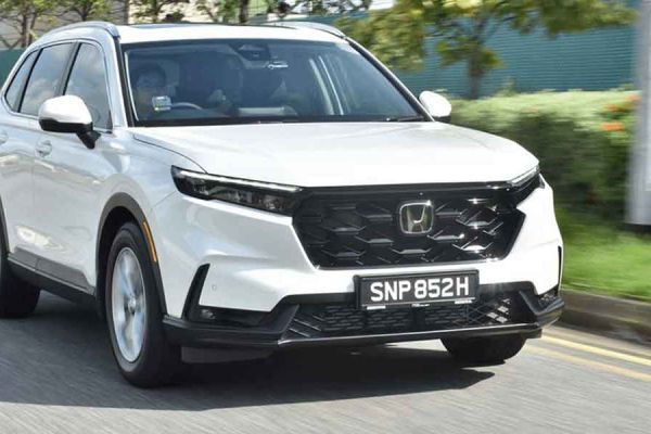 A white Honda SUV driving on a road, with greenery and a blurred background indicating motion, showcasing its sleek front design and dynamic presence.