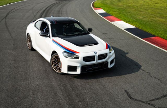 A white BMW sports car with black roof and red-blue stripes, bronze wheels, parked on a racetrack with green grass and curb markings, highlighting its performance and sporty design.