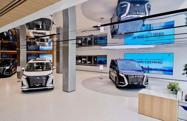 A modern car showroom featuring three luxury MPVs, with sleek designs, large digital screens announcing a grand opening, and a stylish, reflective interior with plants and wooden accents.