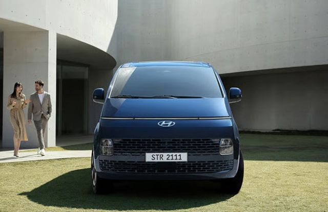 A blue Hyundai minivan with the license plate "STR 2111" parked on a grassy area, with two formally dressed people walking nearby and a modern concrete building in the background.