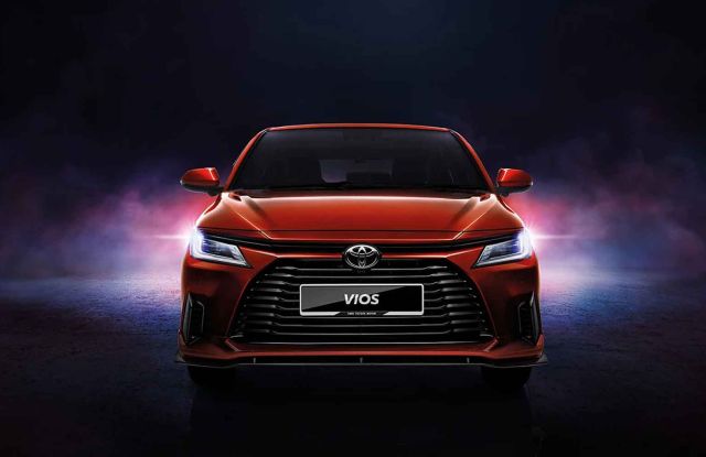 A red Toyota Vios sedan showcased in a dramatic, dark studio setting with colorful lighting effects, emphasizing its bold front design and modern aesthetic.