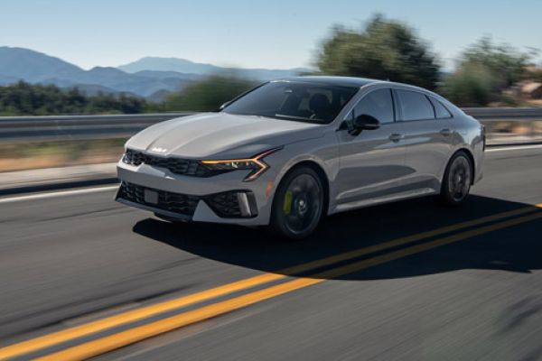 Kia K5 2025 sedan driving on highway (motion blur), mountain/wooded backdrop, yellow road lines.
