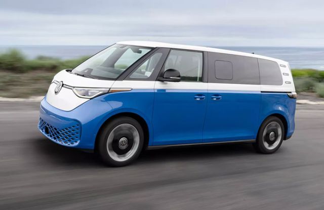 A blue-and-white Volkswagen ID. Buzz van drives along a coastal road, with the ocean and cliffs in the background.