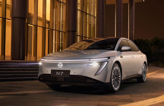 Silver N7 electric sedan parked at night outside a modern building (lit glass walls, columns). The sleek car’s illuminated headlights and "N7" badge are visible; warm lighting and paved ground enhance its contemporary design.
