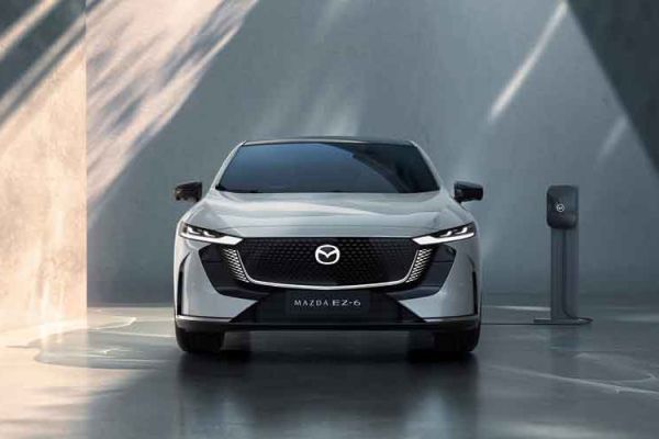 Front view of a silver Mazda EZ-6 electric sedan, charging at a station in a minimalist, futuristic space with dramatic light and shadow on concrete walls.