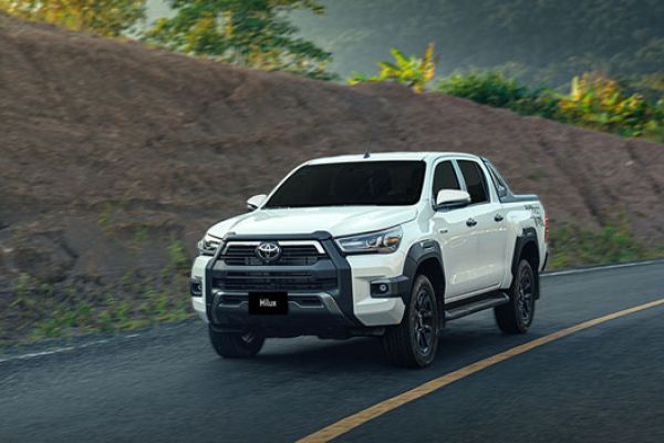A white Toyota Hilux pickup truck driving on a road, with a rugged, green-hilled landscape in the background, showcasing its robust design.