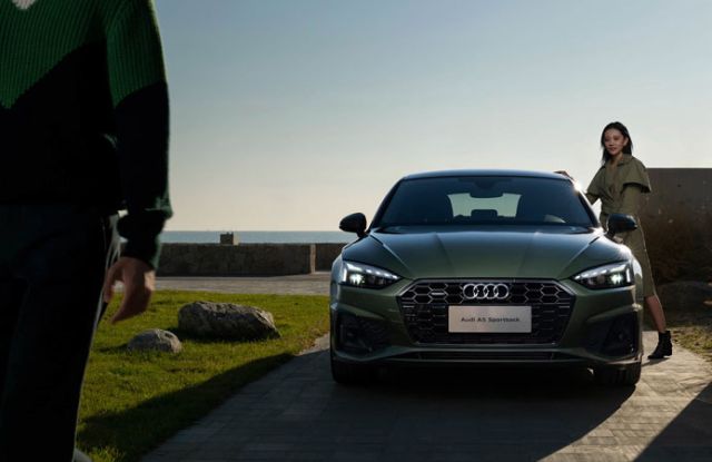Green Audi A5 Sportback (front view): Features Audi logo and badging, parked by a coastal area with two people nearby—showcasing its sleek, premium design in an outdoor setting.