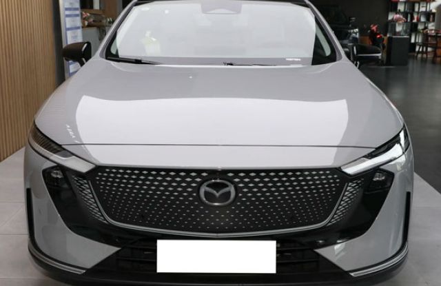 Front view of a light gray Mazda sedan (modern design) in a showroom: Features a textured grille with Mazda logo, slim LED headlights, and sleek black accents. Showroom interior (furniture, displays) visible in the background.