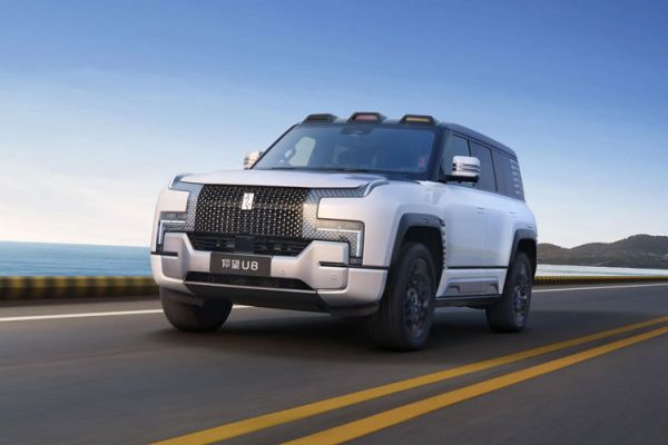 A white 仰望 U8 SUV drives on a coastal highway. Its bold grille, sleek body, and black accents stand out against the blue sea, distant hills, and clear sky, blending rugged style with modern design.